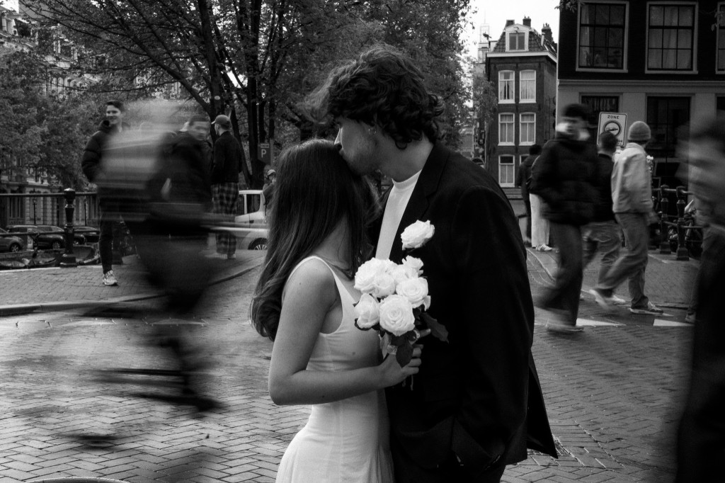 Groom kissing bride's head on a cobblestone street, white roses, motion-blurred crowd