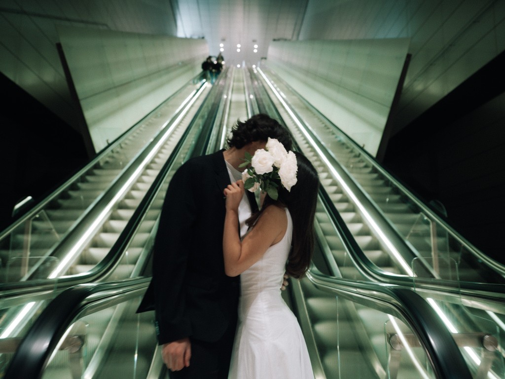 Bride and groom on an escalator, faces hidden behind a bouquet of white roses