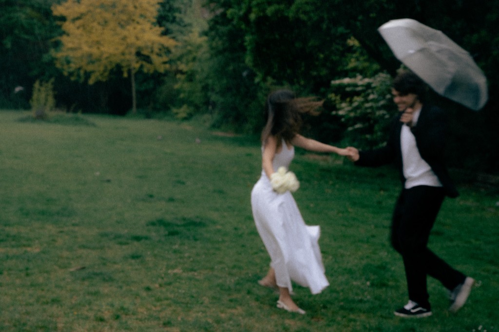 Bride spinning in a white dress beside groom with umbrella in a green garden