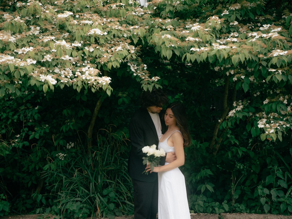 Bride and groom in an intimate embrace before a wall of white flowering greenery