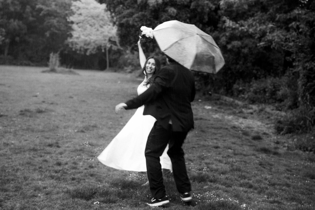 Bride and groom dancing in the rain in a field, umbrella overhead, black and white