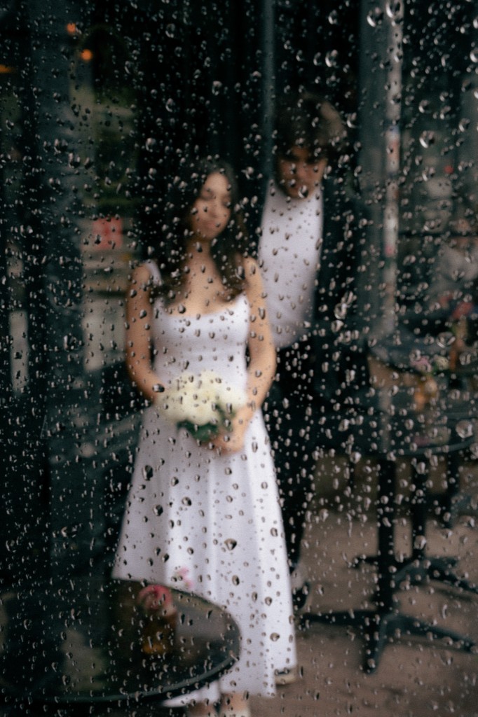 Bride and groom seen through a rain-streaked window, she holds white flowers