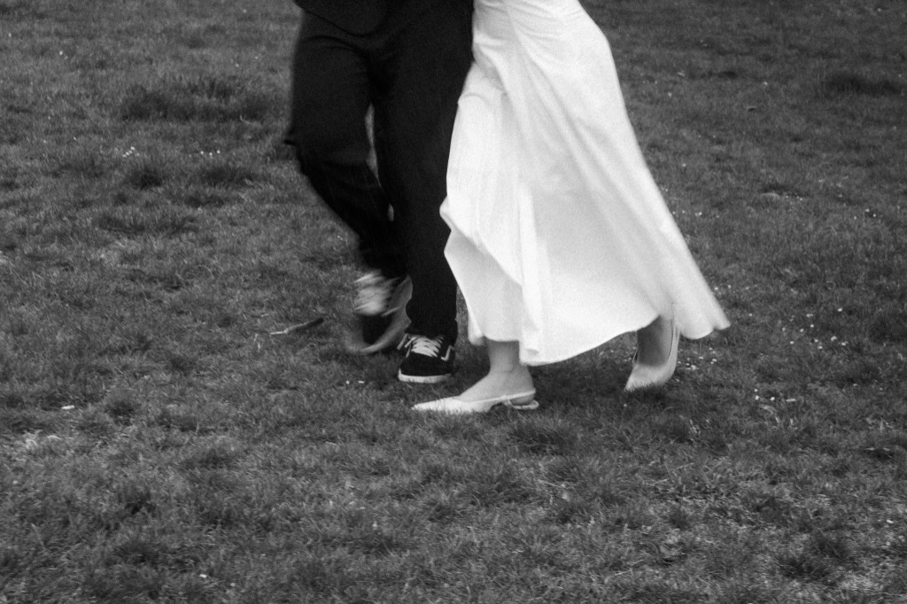Bride in white heels and groom in sneakers walking across grass, black and white